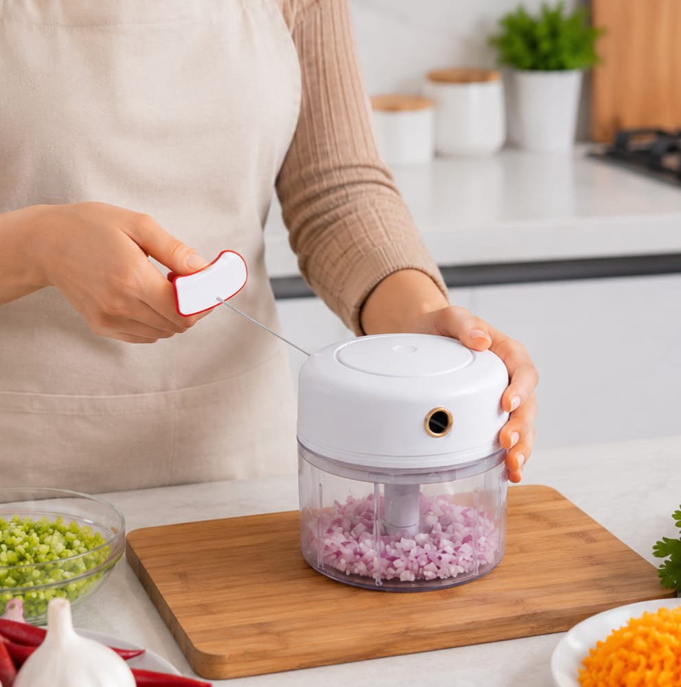 A person in a beige apron uses a white and clear electric food chopper to shred red onion on a wooden cutting board in a bright kitchen.