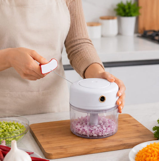 A person in a beige apron uses a white and clear electric food chopper to shred red onion on a wooden cutting board in a bright kitchen.